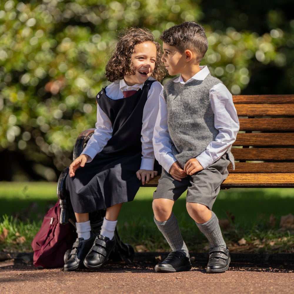 two young kids in school uniform sitting on a bench, both wearing bobux school shoes