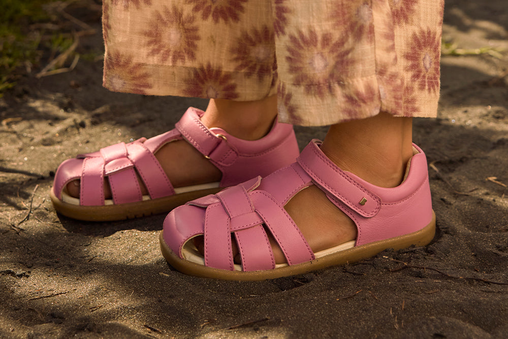Image of a pair of children's feet stood on an earthy floor wearing pink closed-toe Bobux sandals