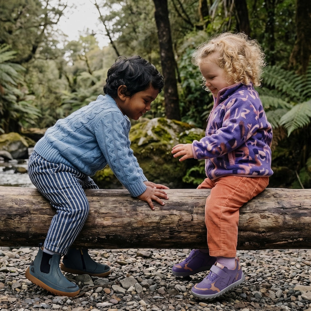 two children sitting on a log in a forest they are wearing bobux barefoot alpha shoes