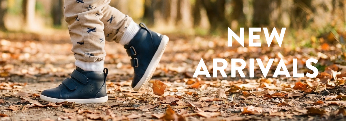 A child's legs wearing beige patterned pants and navy blue ankle boots walking on a forest floor covered in fallen leaves, with the text 'NEW ARRIVALS' overlaid on the right.