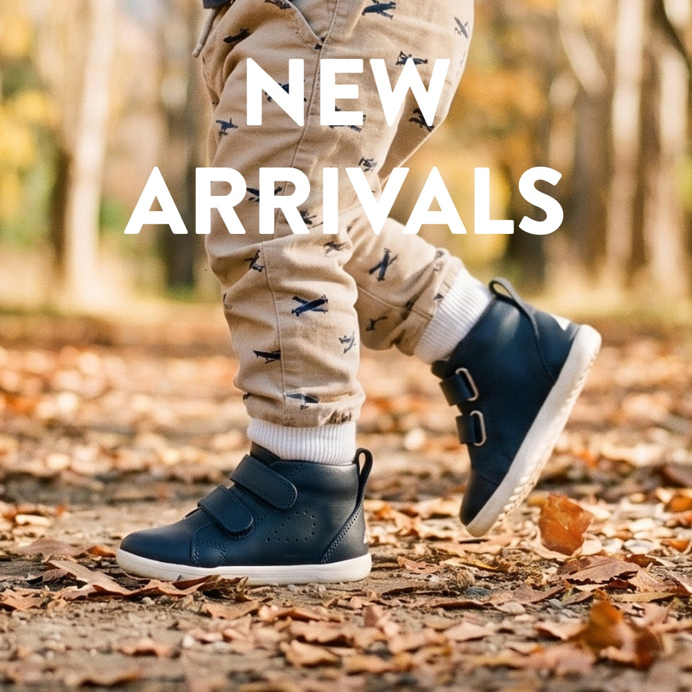 A child's lower legs wearing beige patterned pants and dark navy blue high-top slip-on shoes with white soles, walking on a forest path covered in fallen leaves. Large white text overlay reads 'NEW ARRIVALS'.