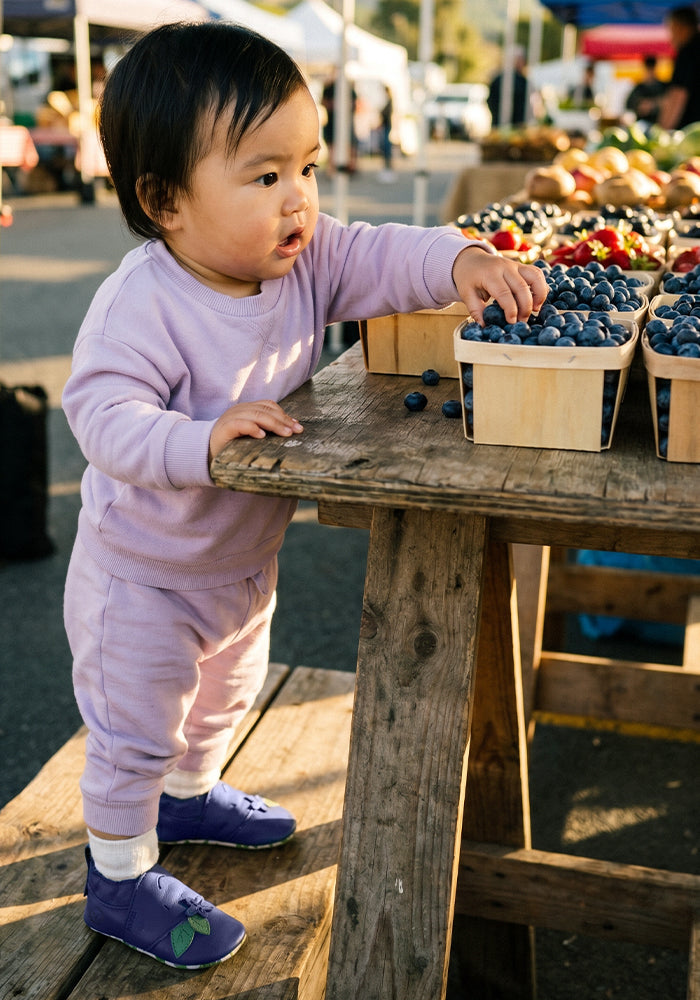 A toddler in a light purple sweatshirt and matching trousers stands at a wooden market table, reaching for blueberries in baskets. The child is wearing purple Bobux shoes and white socks, with an outdoor farmers market scene in the background.